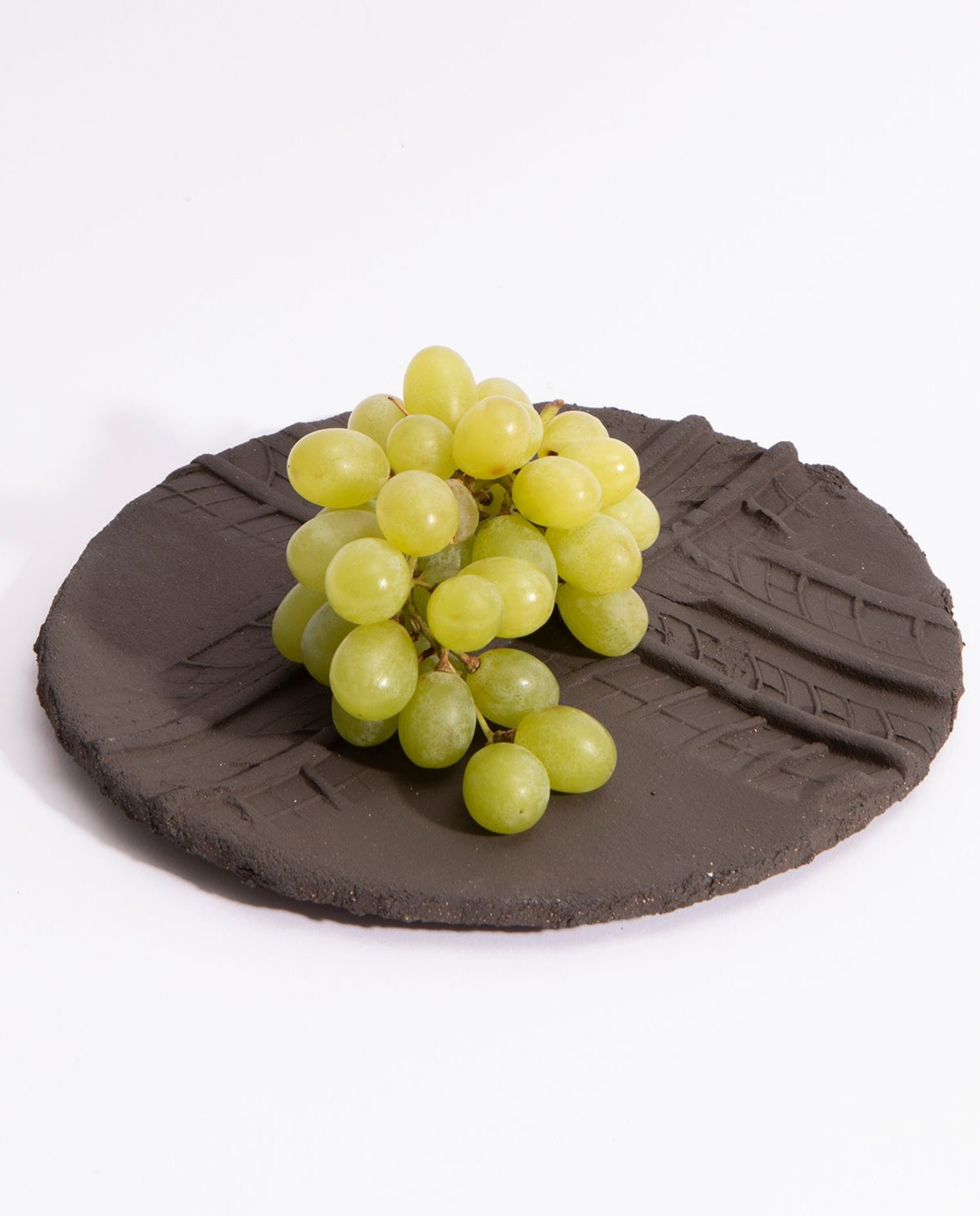 Assiette en grès noire brute, avec deux empreintes de pneu qui se croisent en son milieu. Une grappe de raisin est posé dessus.