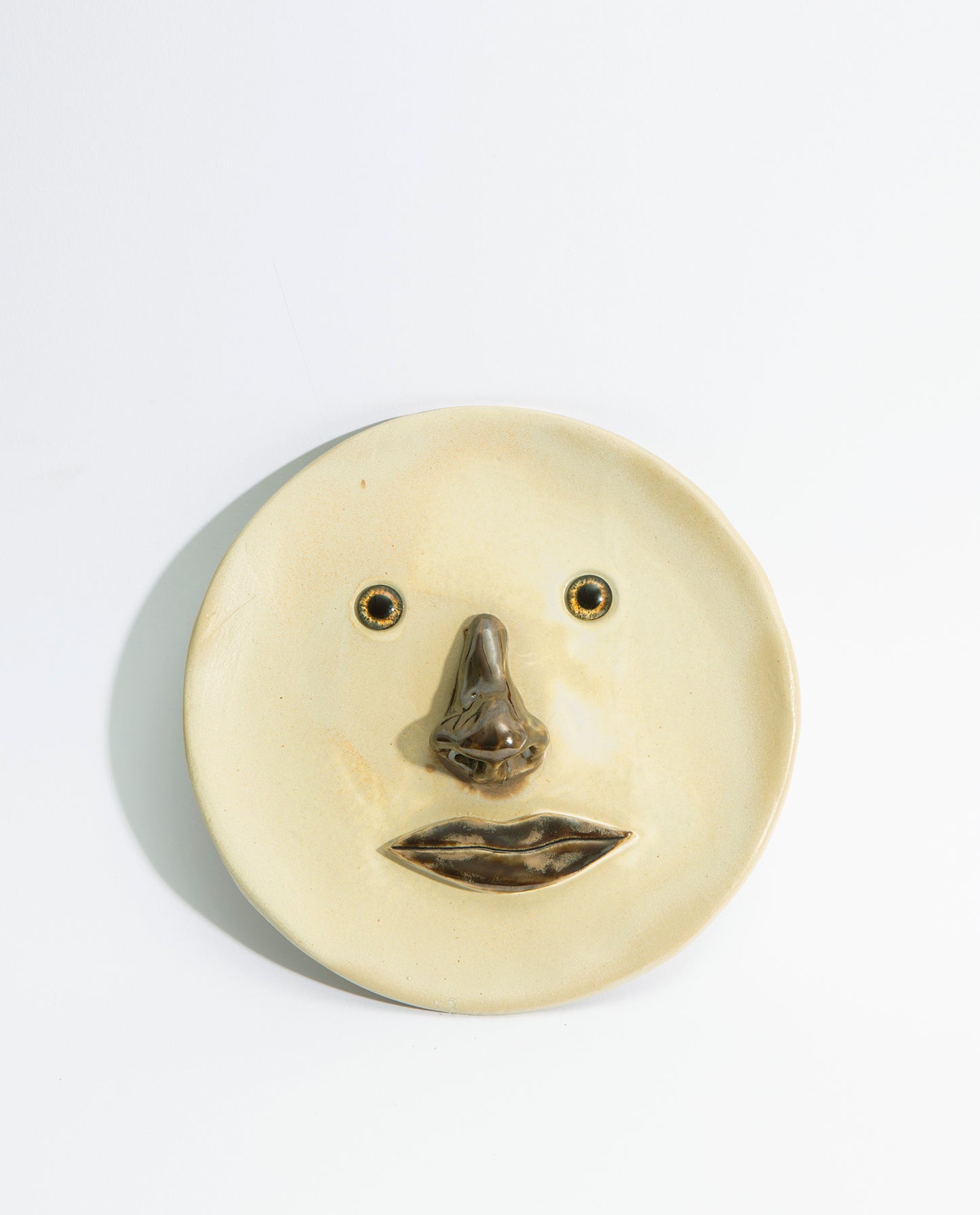 Assiette en grès émaillé beige, avec un nez et une bouche en volume de couleur marron et deux yeux en verre.