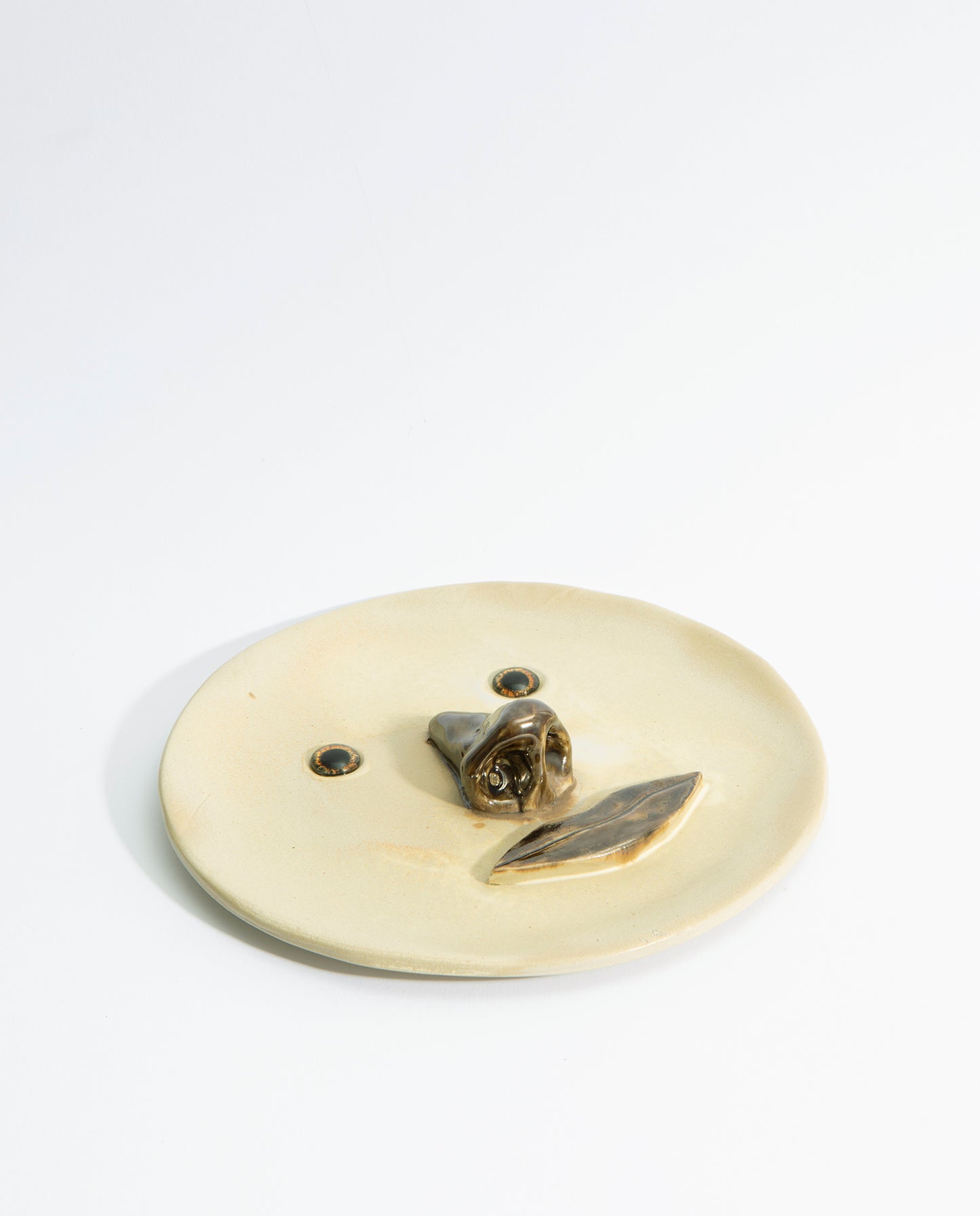 Assiette en grès émaillé beige, avec un nez et une bouche en volume de couleur marron et deux yeux en verre.