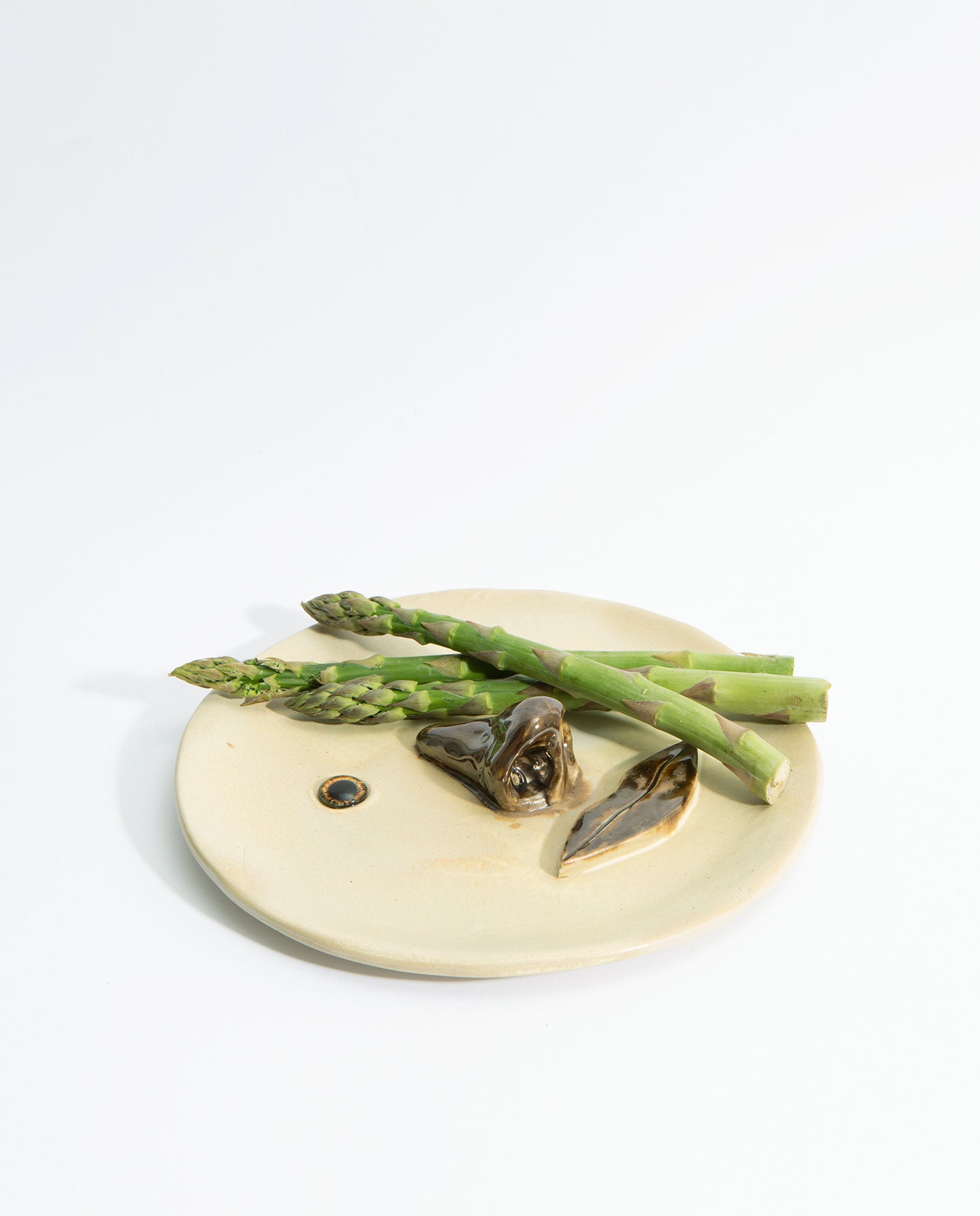 Assiette en grès émaillé beige, avec un nez et une bouche en volume de couleur marron et deux yeux en verre. Trois asperges vertes sont posées dessus.