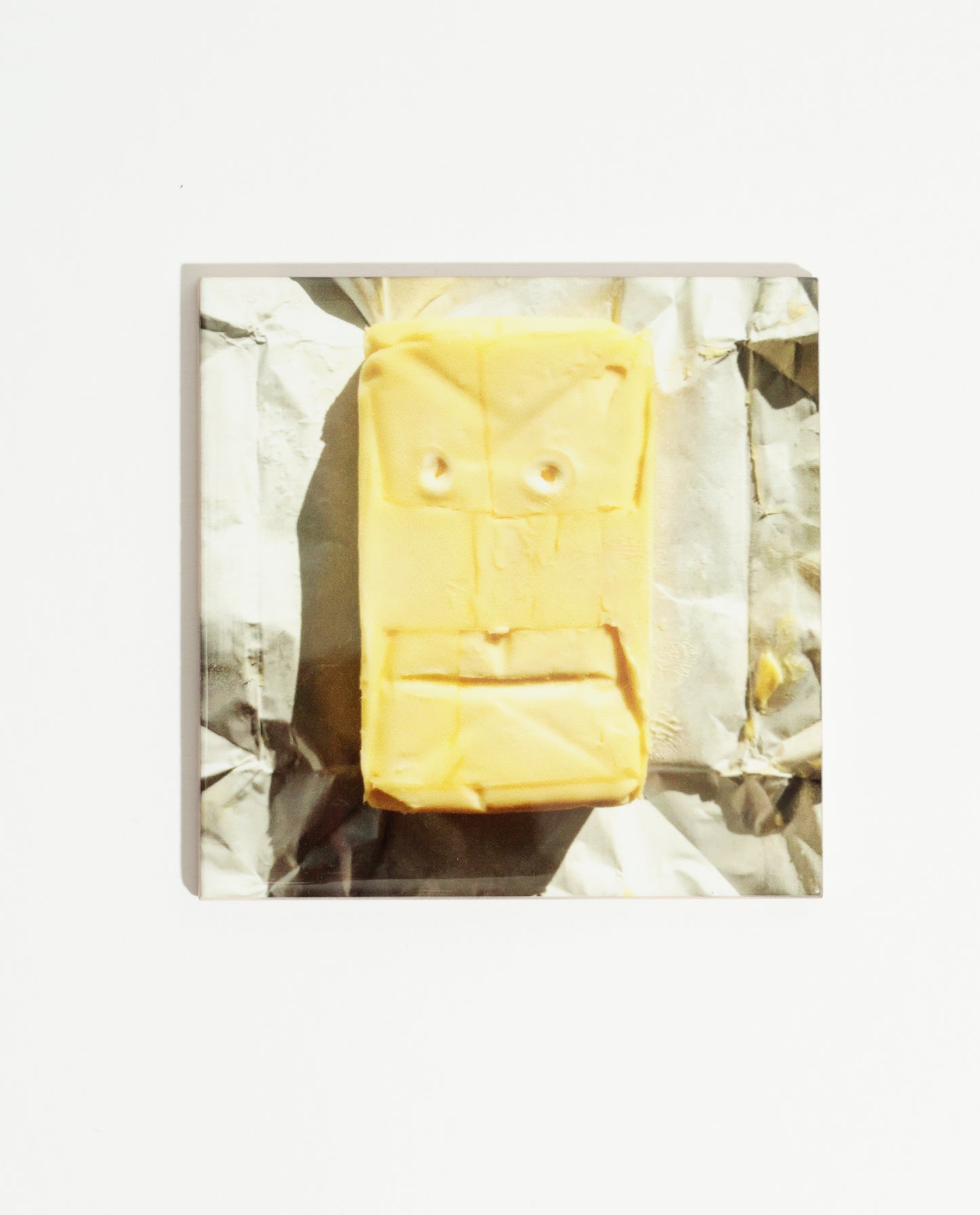 Carreau de faïence imprimée d'une photo de l'artiste Olaf Breuning représentant un visage dans une plaque de beurre. Celui-ci peut être intégré à une faïence de cuisine ou accroché dans la maison.